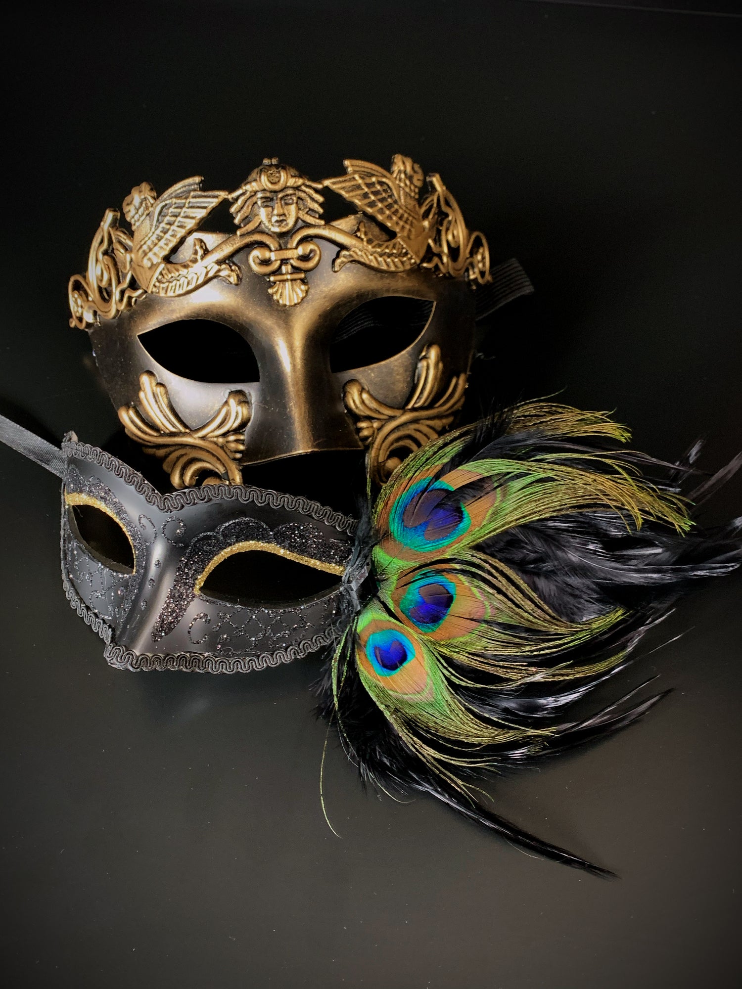 Couples masquerade mask set with a gold gladiator mask for the men and a black peacock mask with gold glitter eyes for the women.