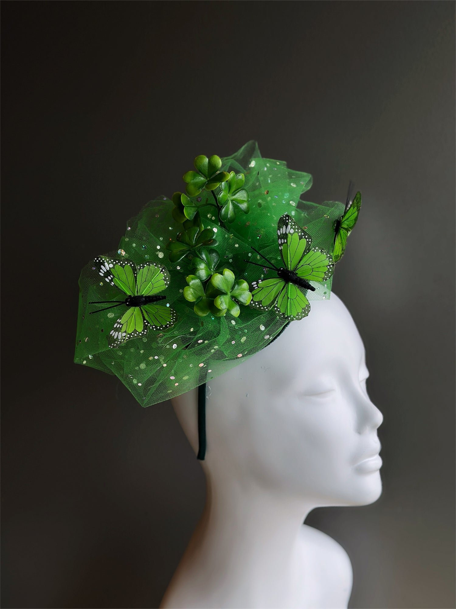 Green fascinator hat with four-leaf clovers, butterflies, and glittering tulle, designed for festive occasions and year-round elegance.