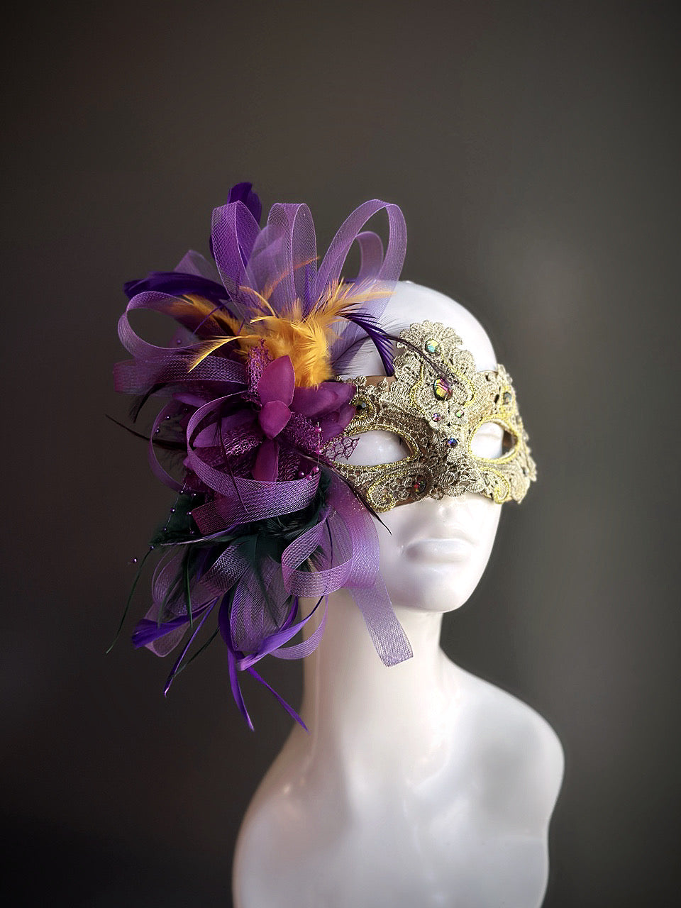 Gold lace masquerade mask with purple feathers, ribbons, and decorative accents displayed on a mannequin head.

