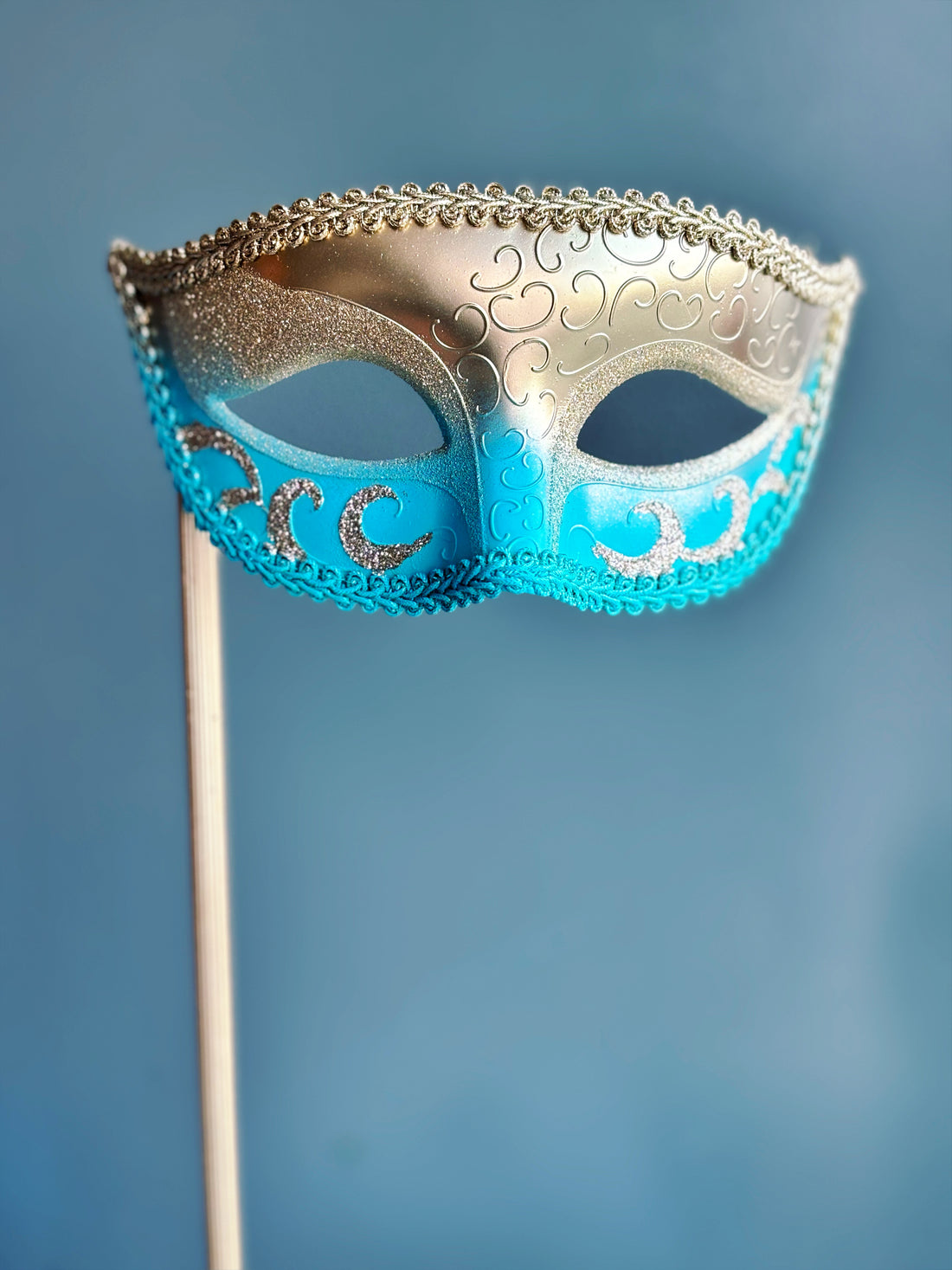 Handheld blue and gold Venetian mask with intricate scrollwork and a decorative stick handle for formal events.