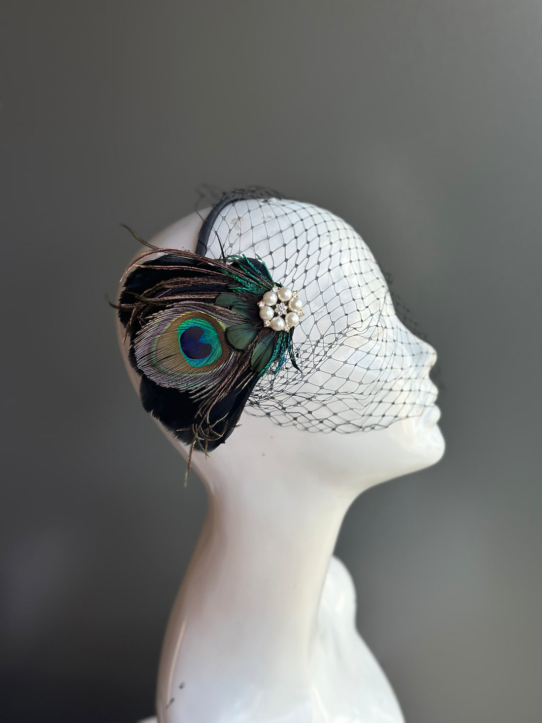 Peacock feather fascinator with a birdcage veil, adorned with pearls and rhinestones, ideal for weddings and elegant events.

