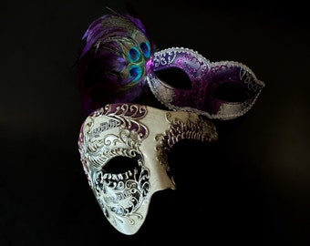 Purple and silver Venetian-style couple masks, one with ornate detailing and the other accented with a vibrant peacock feather, perfect for formal events.