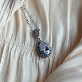 Silver necklace with a teardrop-cut crystal surrounded by a halo of sparkling stones, displayed on ruched satin fabric.