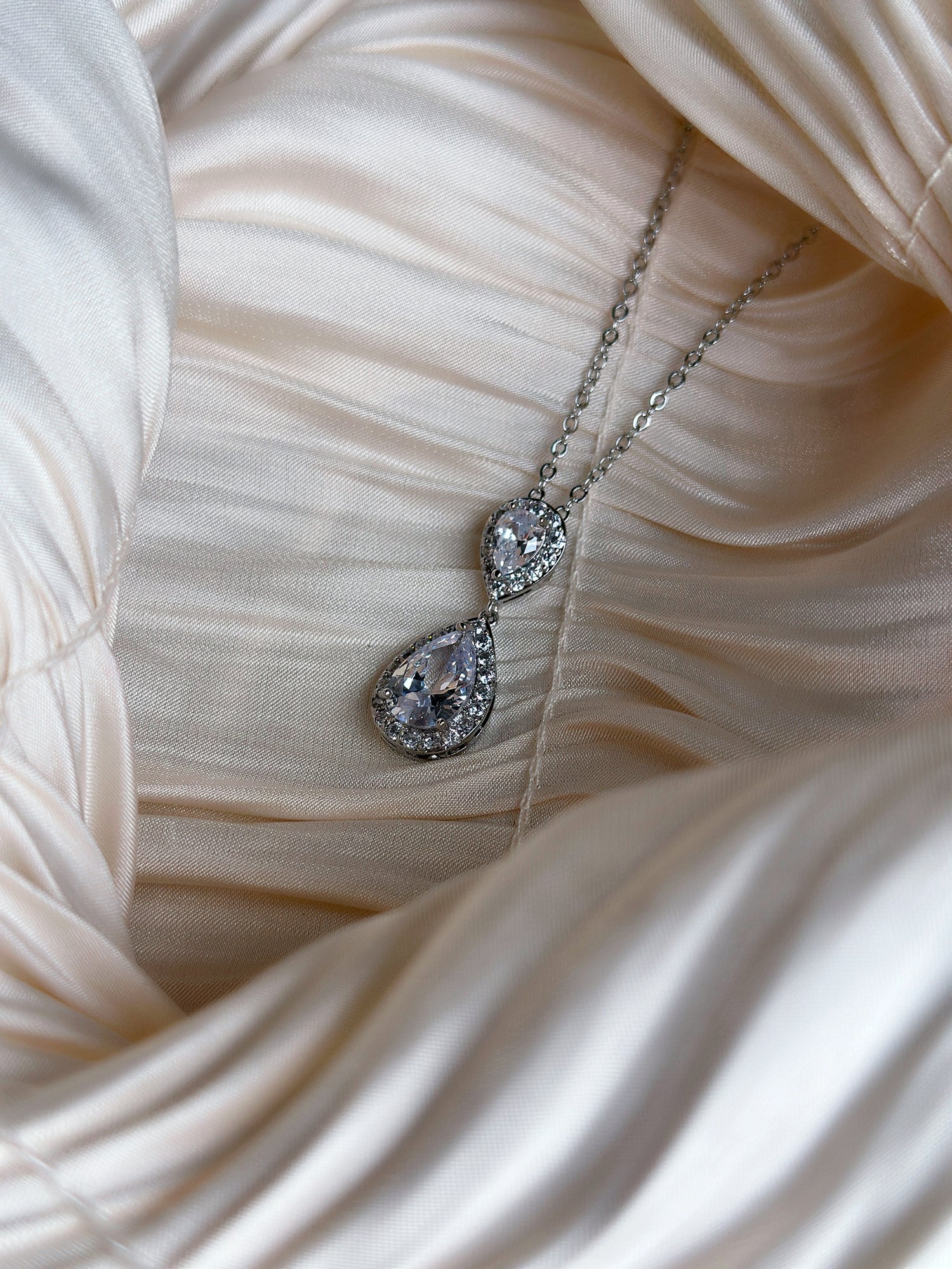 Silver necklace with two teardrop-cut crystals surrounded by a halo of sparkling stones, displayed on ruched satin fabric