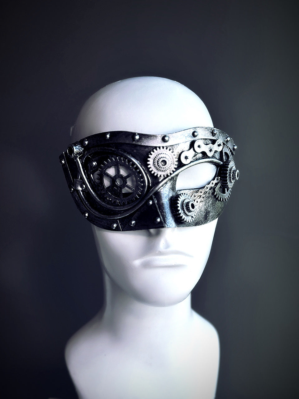 Silver steampunk masquerade mask with gears, chain details, and industrial elements displayed on a mannequin head.

