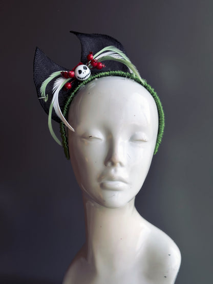 A gothic-inspired headband with black bat wings, a skull centerpiece, red berries, and green feather accents. Ideal for spooky holiday celebrations.